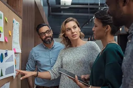 equipe com quatro pessoas, duas mulheres e dois homens, estão em pé e conversando, de frente para uma parede com gráficos expostos. Uma das mulheres está segurando um tablet. A imagem sugere que eles estão em uma discussão de trabalho.