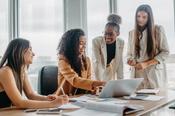 quatro mulheres estão em volta de uma mesa de escritório. Duas estão sentadas e duas estão em pé. Ao centro, uma das moças está utilizando um notebook