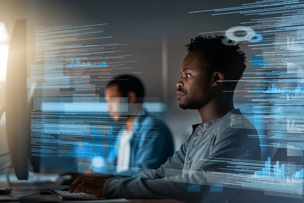 dois rapazes estão sentados frente a telas de computador e em volta deles há hologramas de códigos de programação.