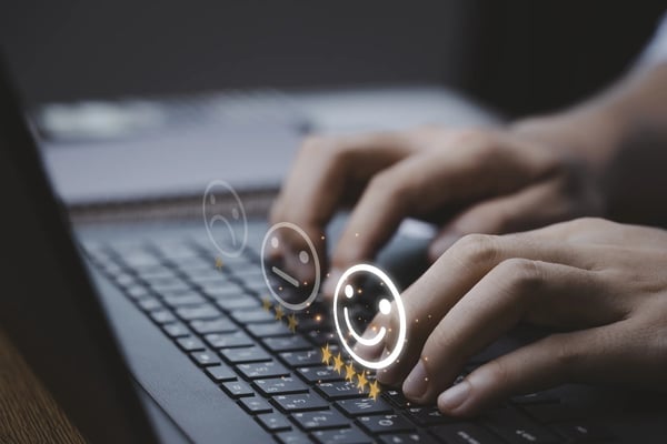 homem digitando em um notebook e em cima do teclado há um holograma com carinhas de feliz, triste e mediano; para avaliação de satisfação.