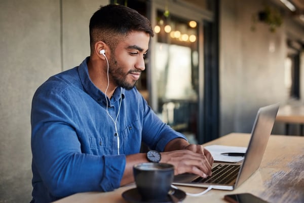 Na imagem, um homem jovem e negro está sentado em frente a um notebook digitando algo. Ele veste uma camisa social azul escuro e sorri. Usa, ainda, fones de ouvido. O tema do texto é North Star Metric e o que é essa métrica aplicada aos negócios.