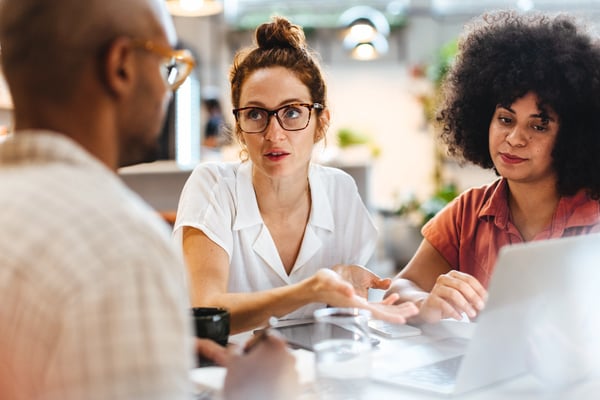 colegas de trabalho em um escritório conversando, no centro uma mulher branca, a sua esquerda uma mulher negra e a direita um homem de costas