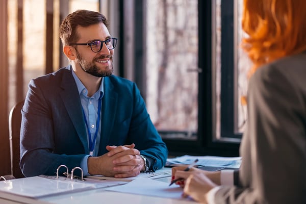 Homem de blazer e óculos sorrindo para uma possível vendedora também de blazer, porém de costas para a imagem