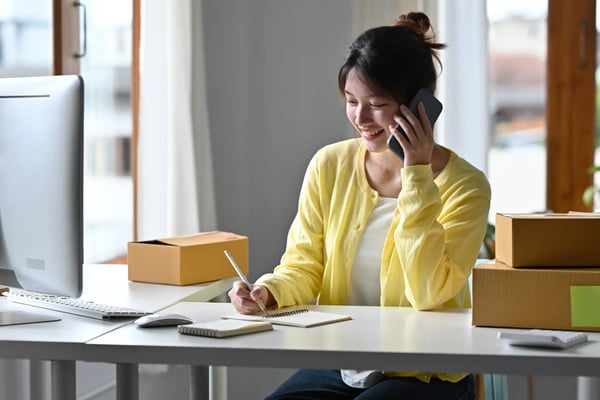 Mulher jovem vestindo amarelo sentada próxima a uma mesa, ela segura um lápis em uma mão e na outra está falando no smartphone