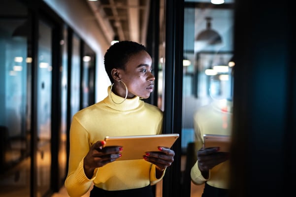 Mulher de pele negra e usando blusa amarela em um ambiente corporativo segurando um tablet em suas mãos.