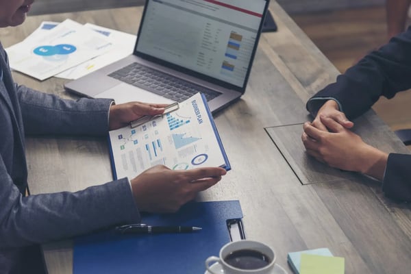 A imagem apresenta duas mãos masculinas, um tablet com gráficos, sobre uma mesa. O tema do artigo é cap table.