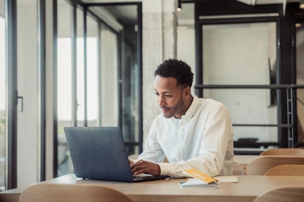 Homem negro sentado em frente a um notebook. No ambiente portas de vidro e uma mesa de madeira.