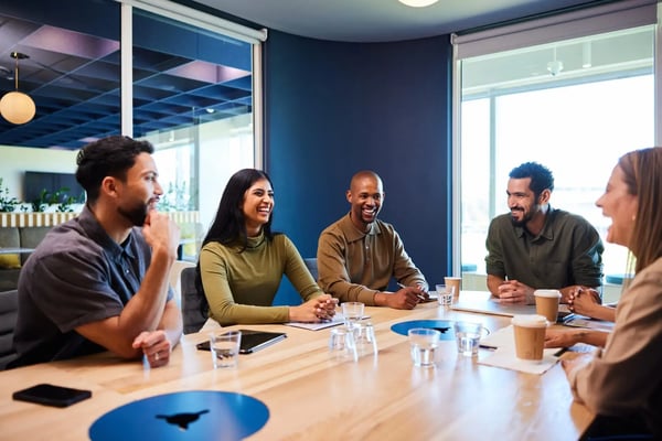 Na imagem, um grupo de cinco pessoas, sendo três homens, todos jovens e multiétnicos, estão sentados ao redor de uma mesa de reuniões em uma empresa. Eles vestem roupas informais e o clima é descontraído. O tema do texto é metodologias de gestão de metas.