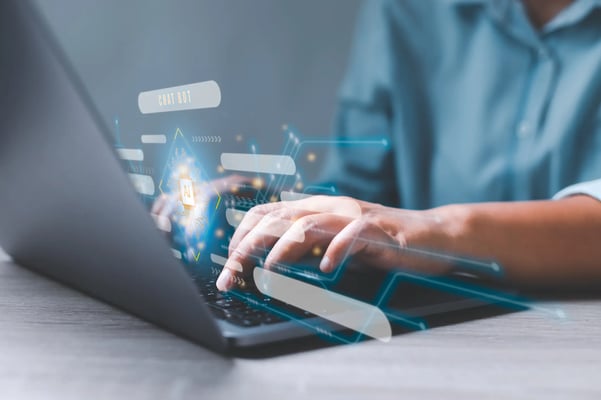 um homem de camisa azul está digitando em seu notebook. A imagem mostra as mãos do rapaz e o notebook. Acima do computador, um holograma.