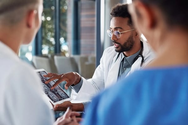 A imagem apresenta um médico, um homem jovem e afro-americano, vestindo jaleco e estetoscópio, que analisa imagens de raio X. Outras duas pessoas de costas acompanham a explicação dele. O tema do artigo é inteligência artificial na saúde.