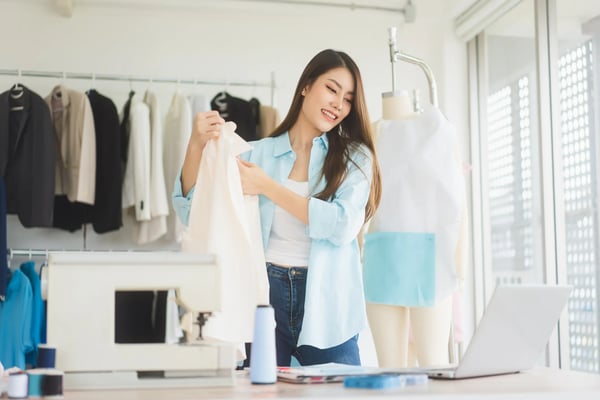 Na imagem, uma mulher de ascendência asiática e jovem, usando roupas casuais, está manuseando uma roupa. Ela tem um computador a sua frente e, no cômodo, há uma arara com roupas, como se fosse uma loja online de roupas. O texto trata sobre o que é DNVB, os negócios nativos digitais.