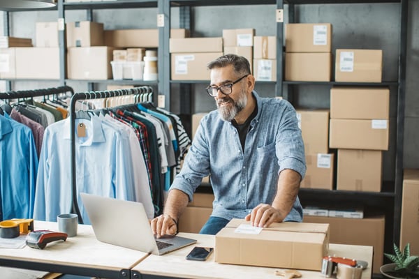 Na imagem, um homem de meia idade branco e vestindo roupas casuais, está em frente a um notebook. Ele está em um depósito com caixas e algumas roupas penduradas em uma arara atrás de si. O texto apresenta o que é DNVB e como criar um negócio desse tipo.