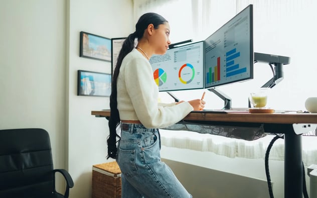 Mulher jovem de pé em frente a dois monitores que contém gráficos.