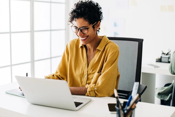 mulher vestindo camisa amarela sentada em seu escritório e sorrindo olhando para seu notebook branco.